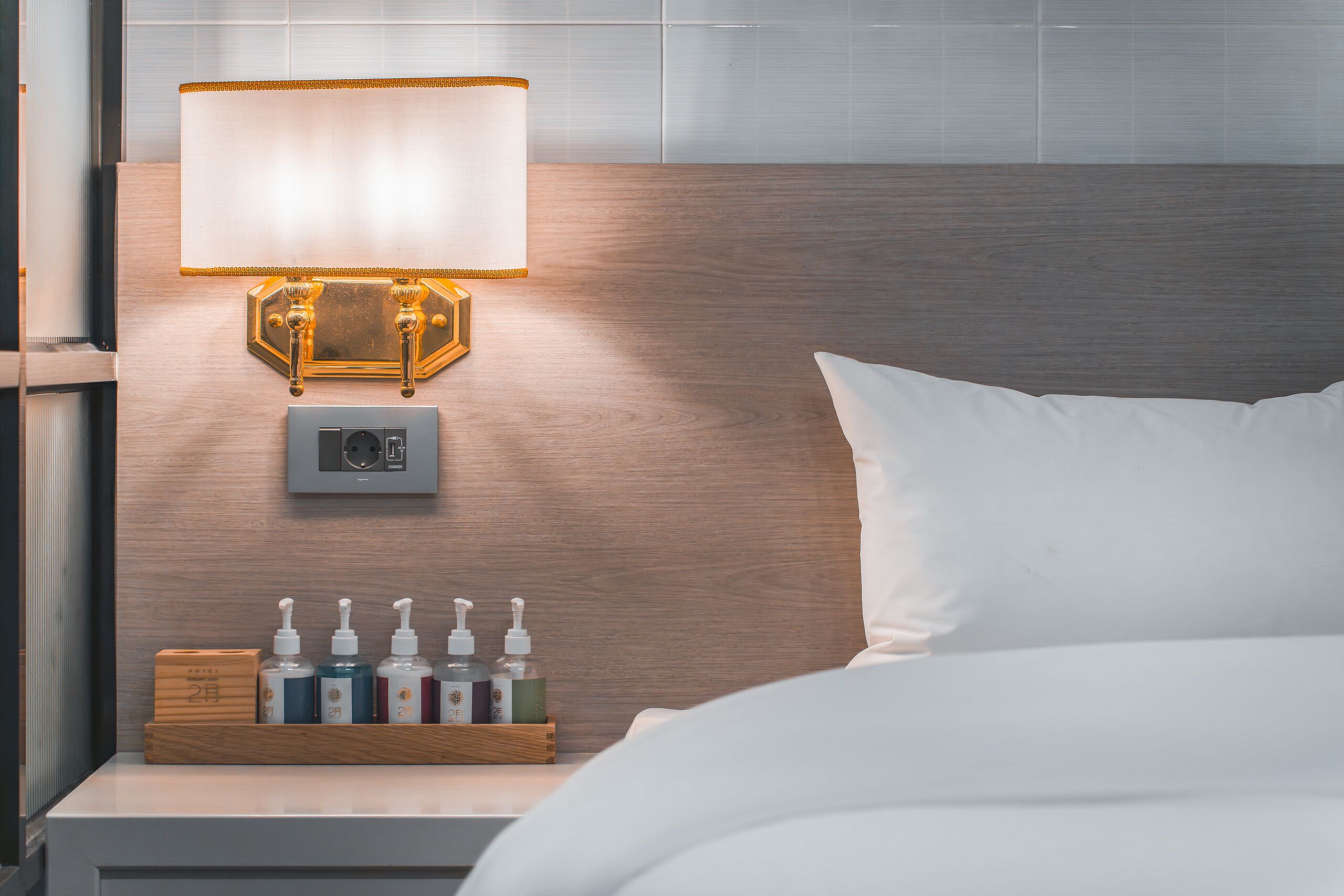 Un lit bien fait, avec un oreiller blanc, une table de chevet sur laquelle se trouve une lampe, et un petit plateau en bois contenant des articles de toilette.