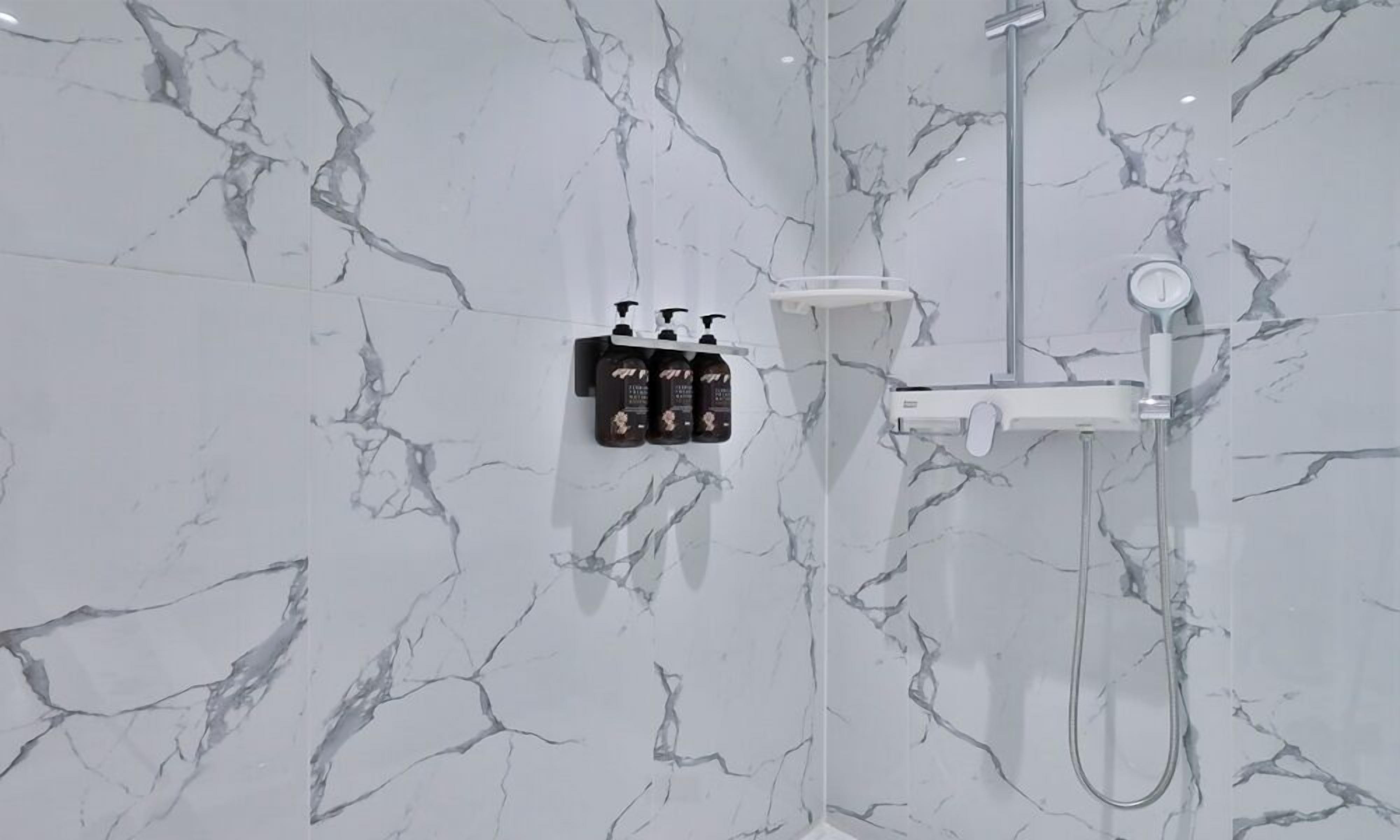 A bathroom with a marble wall, a shower, and a soap dispenser mounted on the wall.