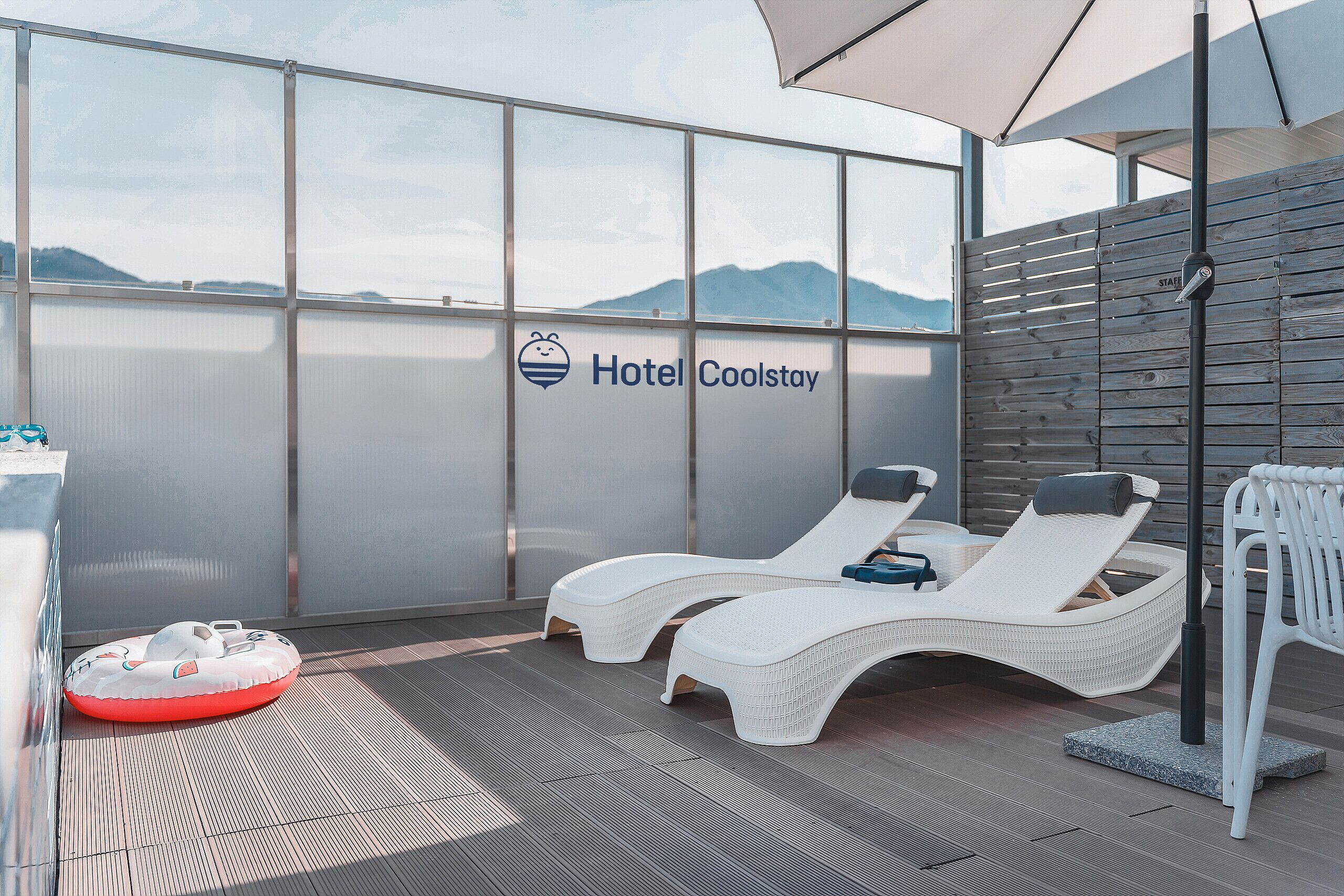 Un balcón iluminado por el sol con vistas a montañas, dos sillones de descanso y una piscina inflable pequeña. Un letrero indica 'Hotel Coolstay'.