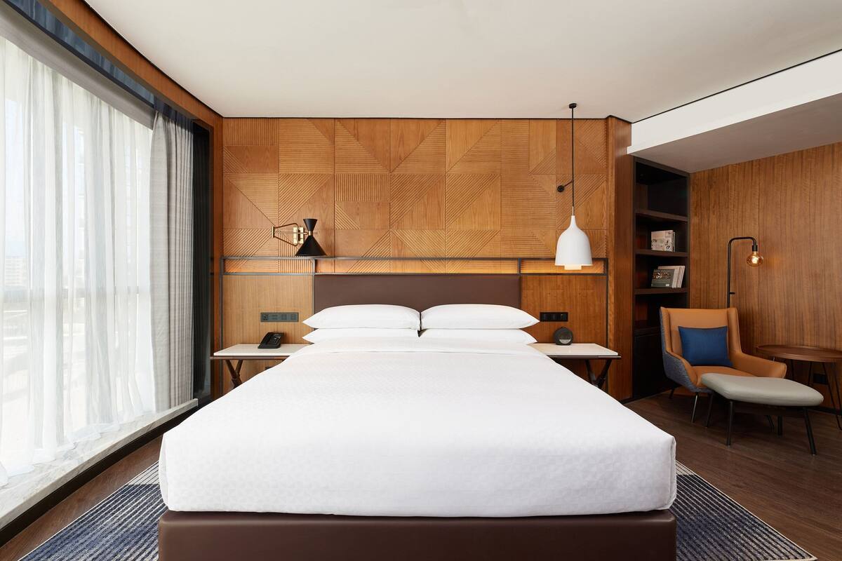 A modern hotel room with a large bed, wooden panel wall, bedside tables, a chair, and a desk.