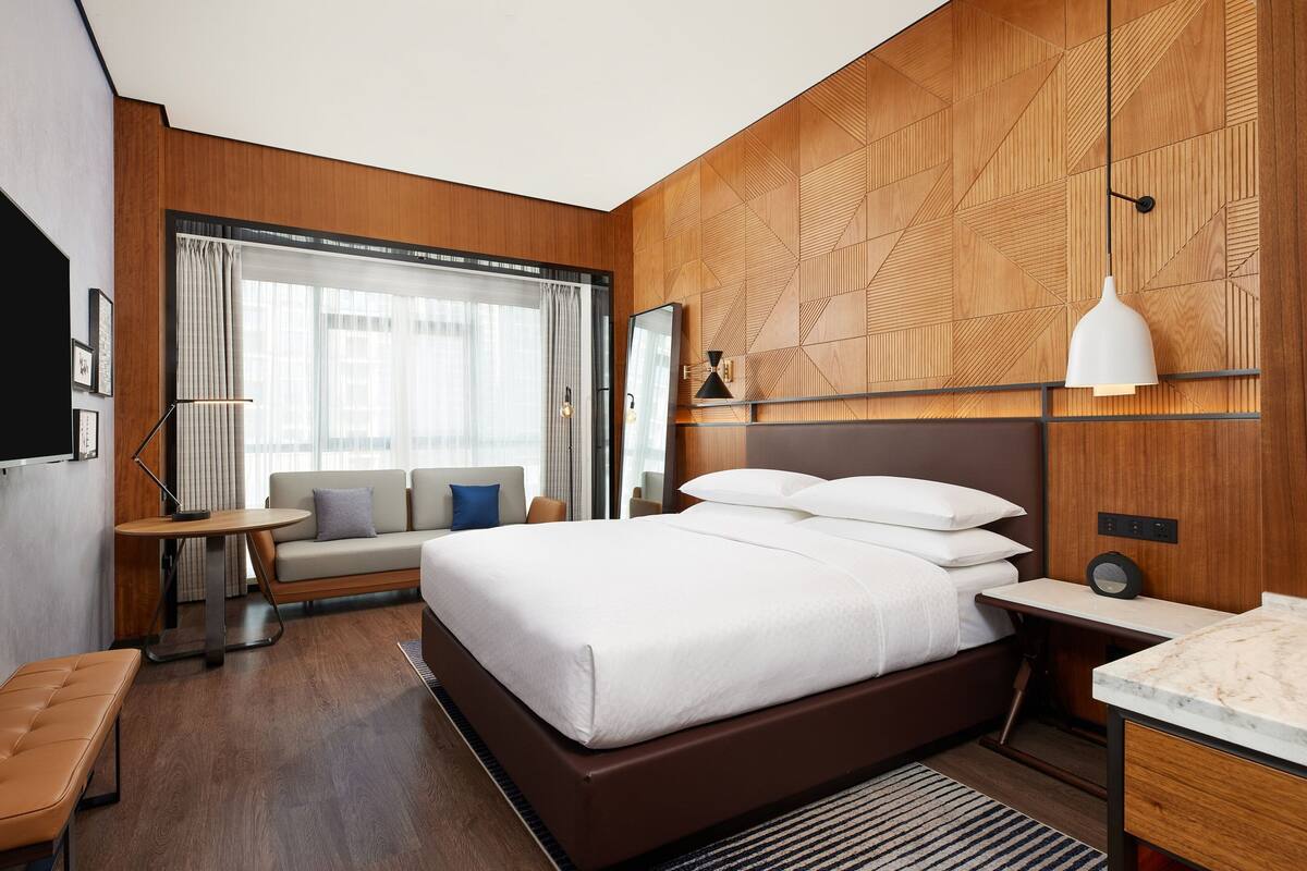 A modern hotel room with a large bed, a seating area, a desk, and a large window with curtains.