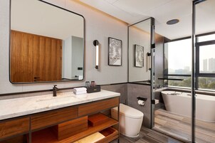 A modern bathroom with a large mirror, wooden vanity, a bathtub, and a city view through a large window.