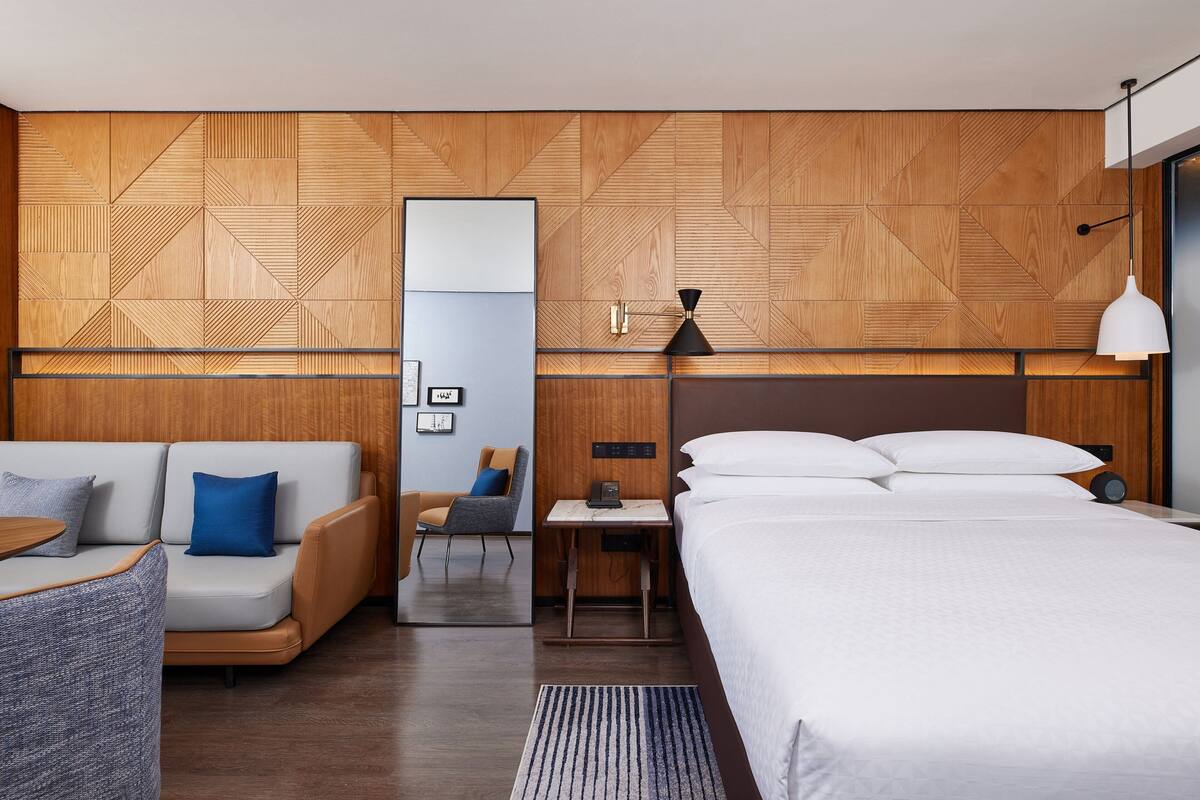 A modern hotel room with a wooden accent wall, a sofa, a bed, a desk, and a chair.