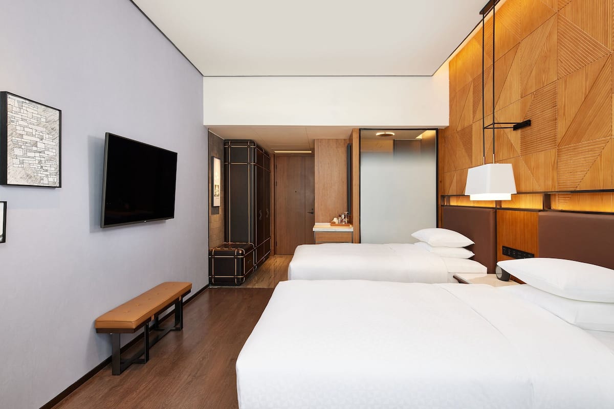 A hotel room with two beds, a bench, a TV, a wooden wall panel, and a mirror.