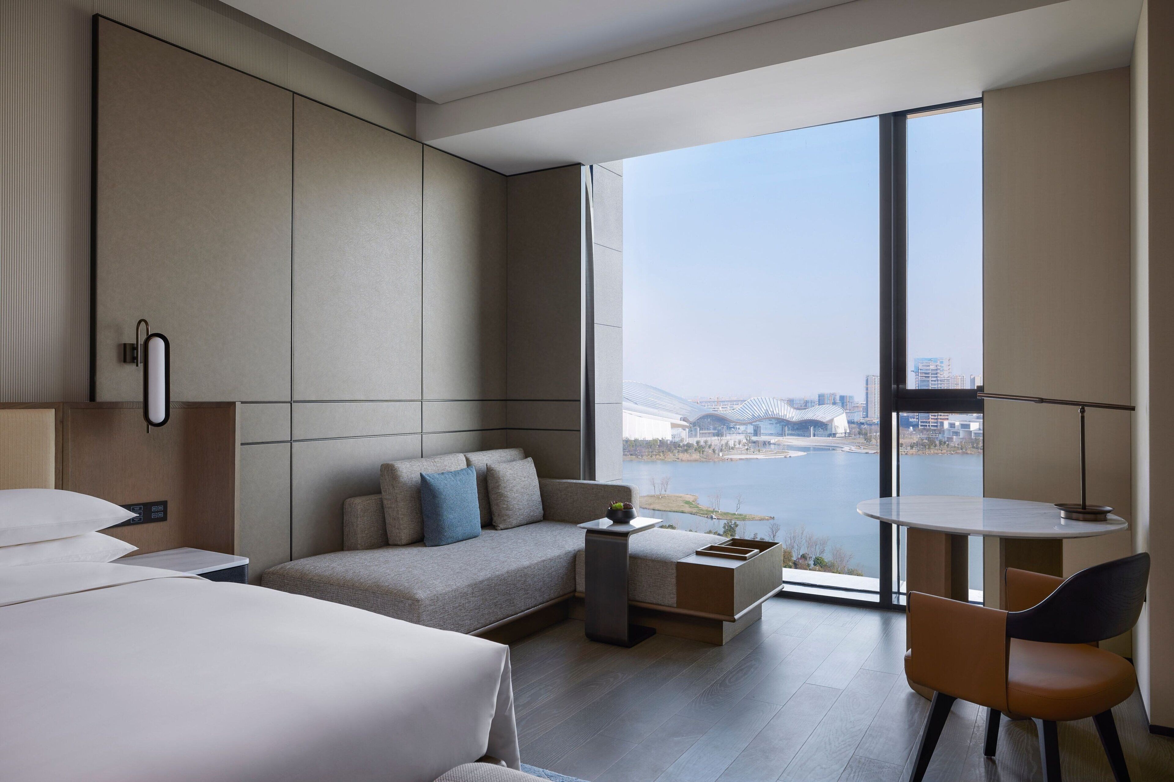 A modern hotel room with a large window, a sofa, a desk, and a chair.