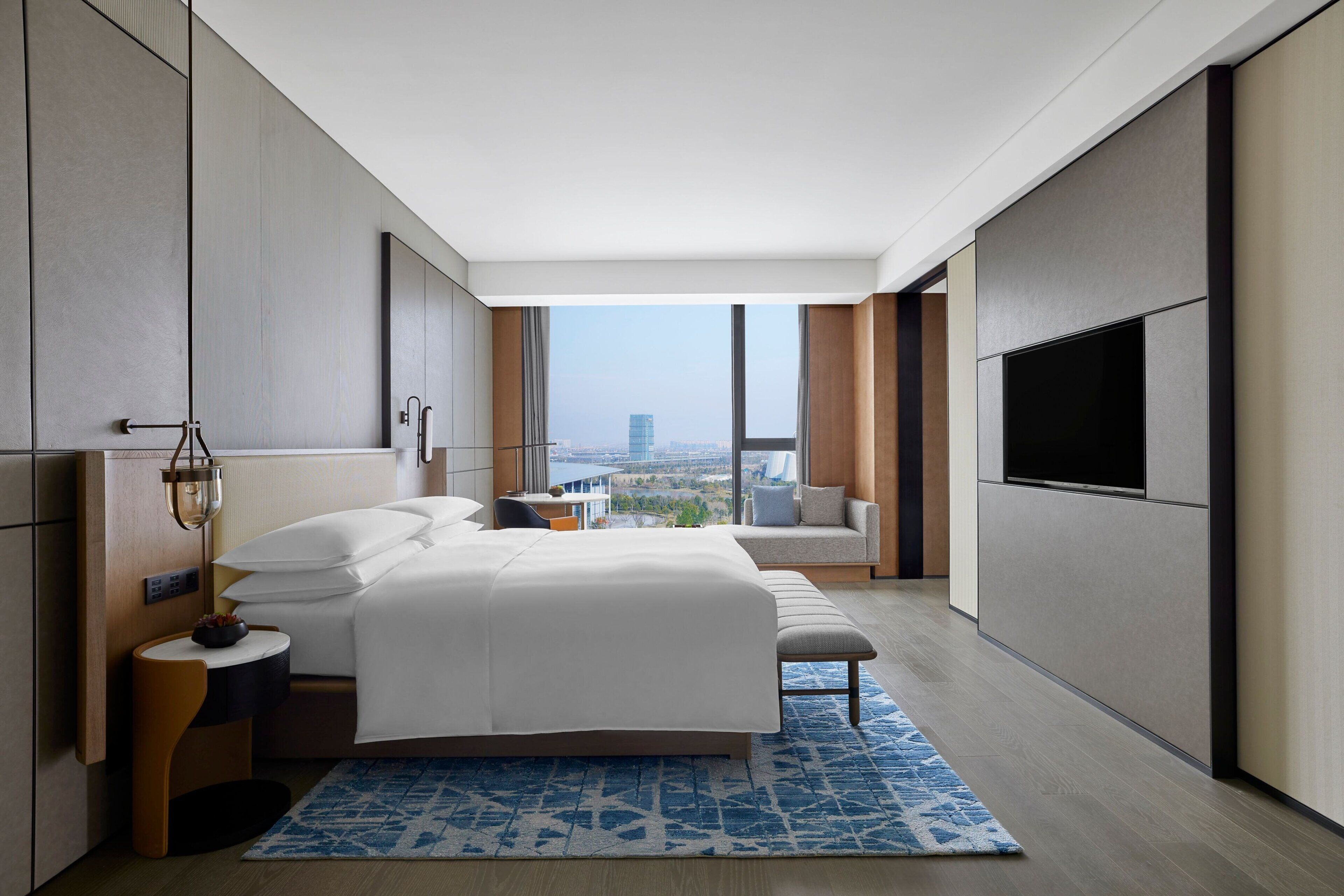 A modern hotel room with a large bed, a TV, a view of the city, and a small seating area.