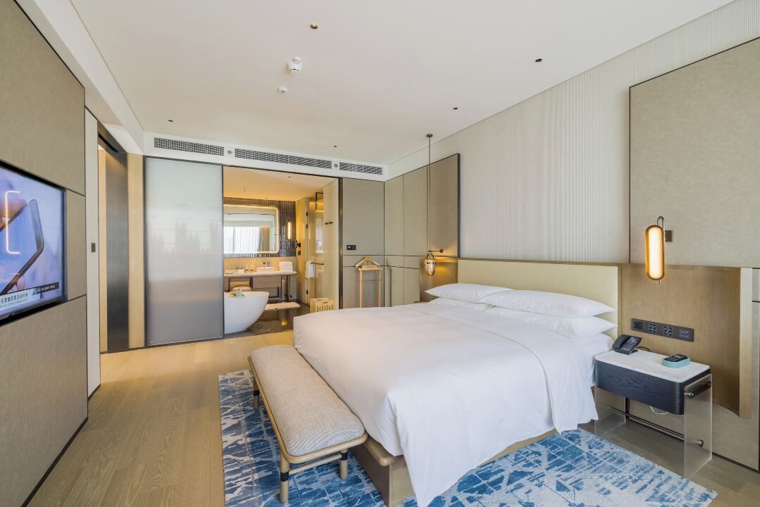 Una camera d'albergo moderna con un letto grande, una panca, un comodino, una TV e un bagno visibile attraverso una porta aperta.