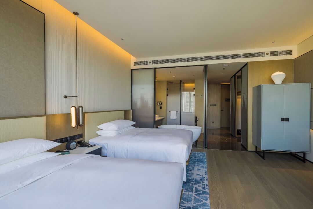 A hotel room with three beds, a large mirror, a wardrobe, and a bathroom visible through a glass door.