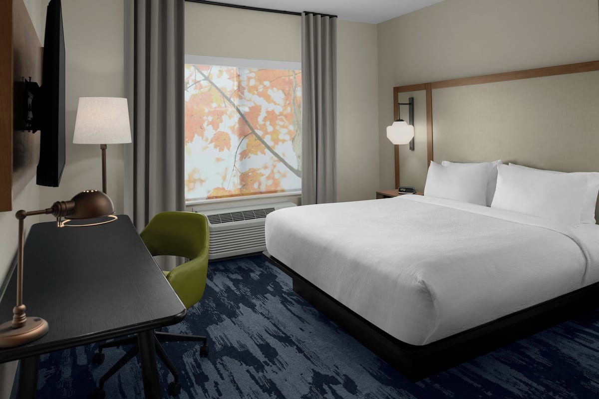 Ein Hotelzimmer mit einem großen Bett, einem Schreibtisch mit Lampe, einem Sessel und einem Fenster mit Vorhängen.