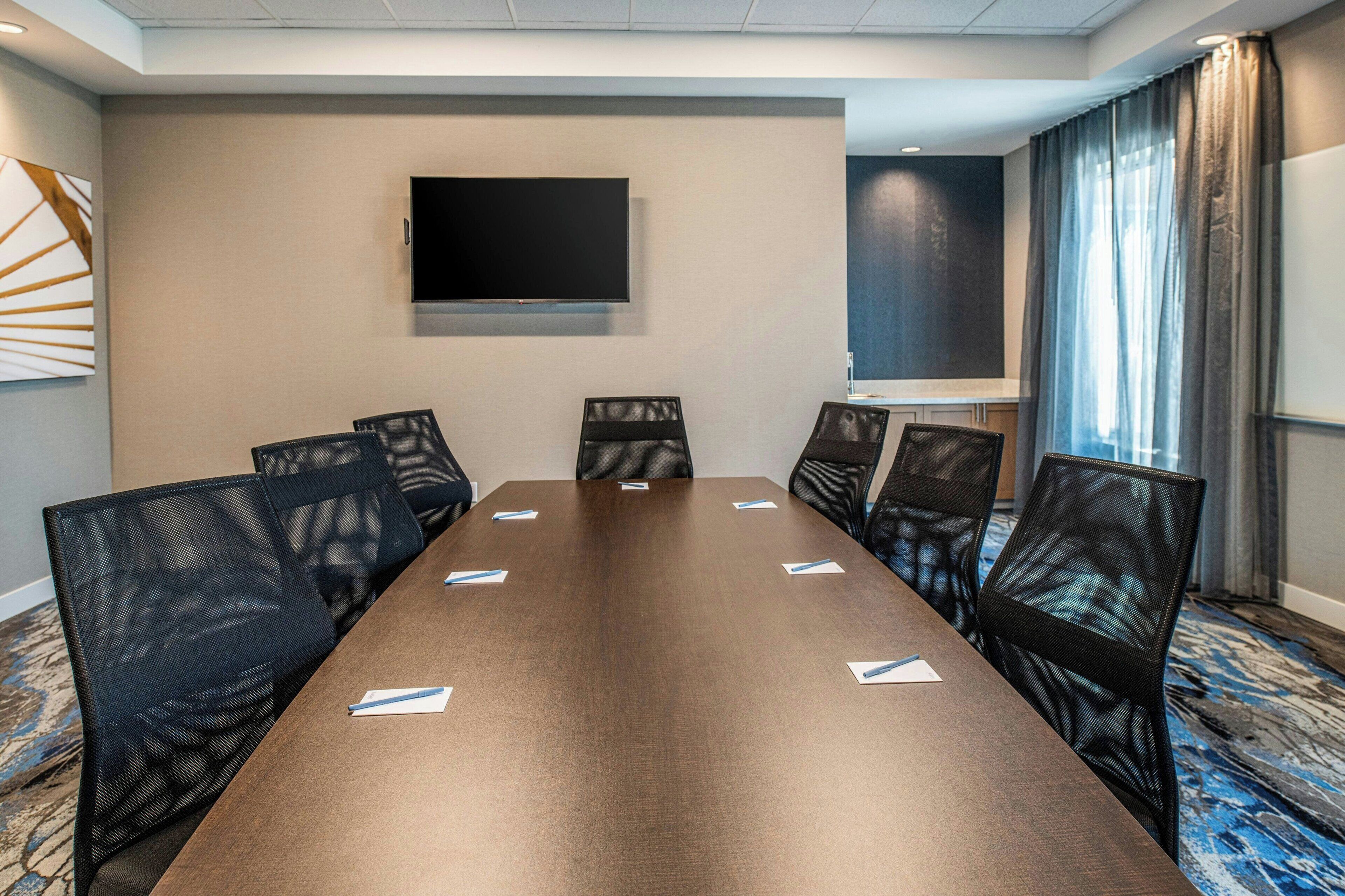 A conference room with a long table, six chairs, a flat-screen TV, and a window with curtains.