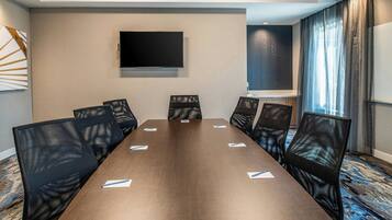 A conference room with a long table, six chairs, a flat-screen TV, and a window with curtains.