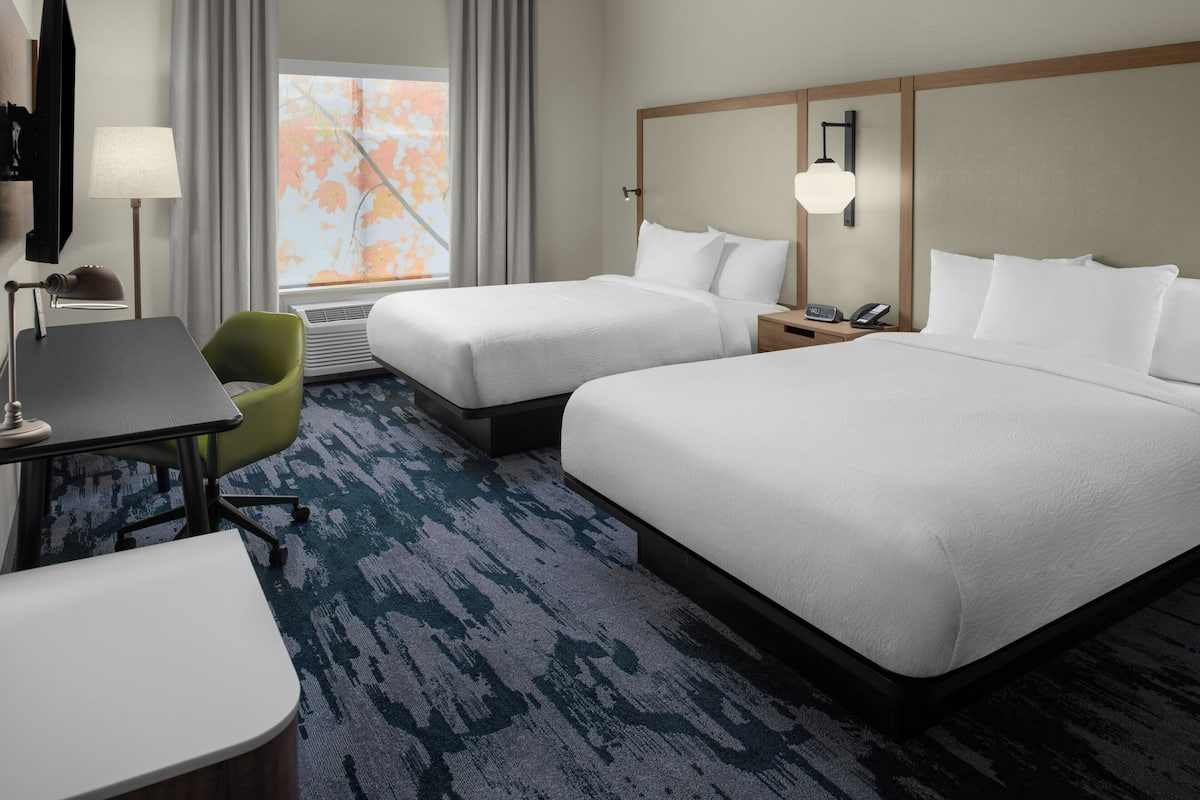 Ein Hotelzimmer mit zwei Betten, einem Schreibtisch, einem Sessel, einer Lampe, einem Telefon und einem Fenster mit Vorhängen.