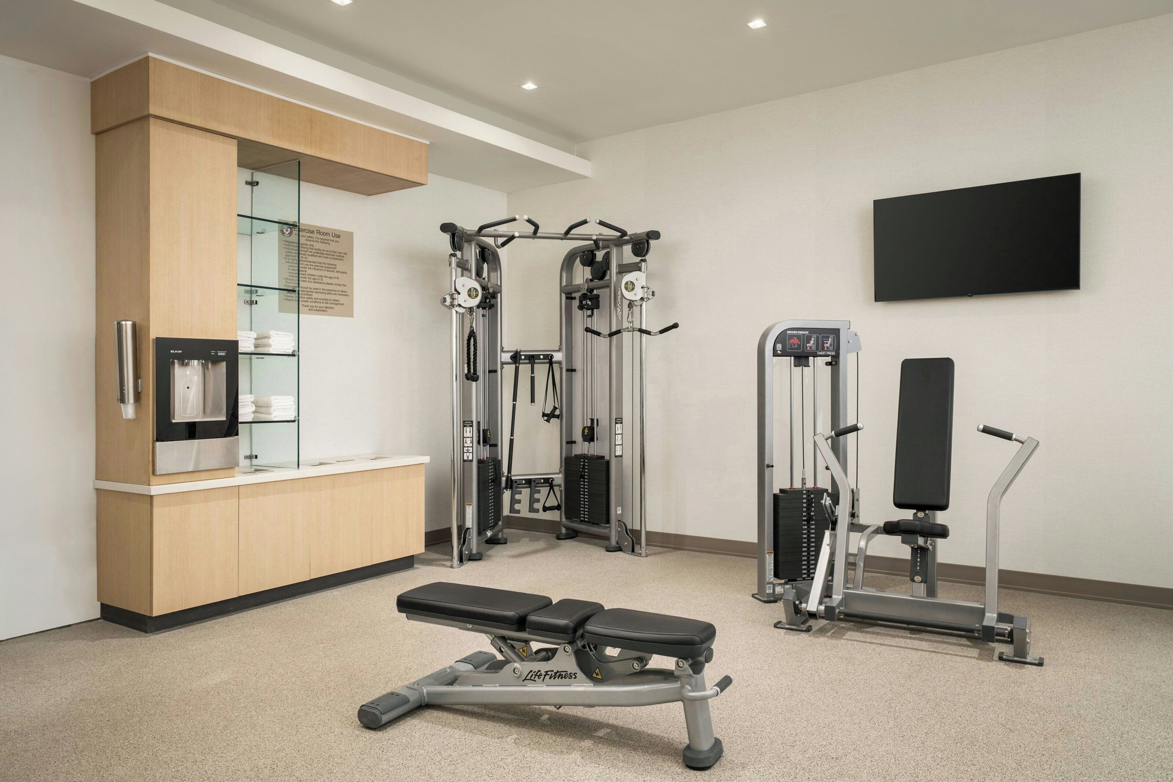 A fitness room with a bench, weight machines, and a wall-mounted TV.