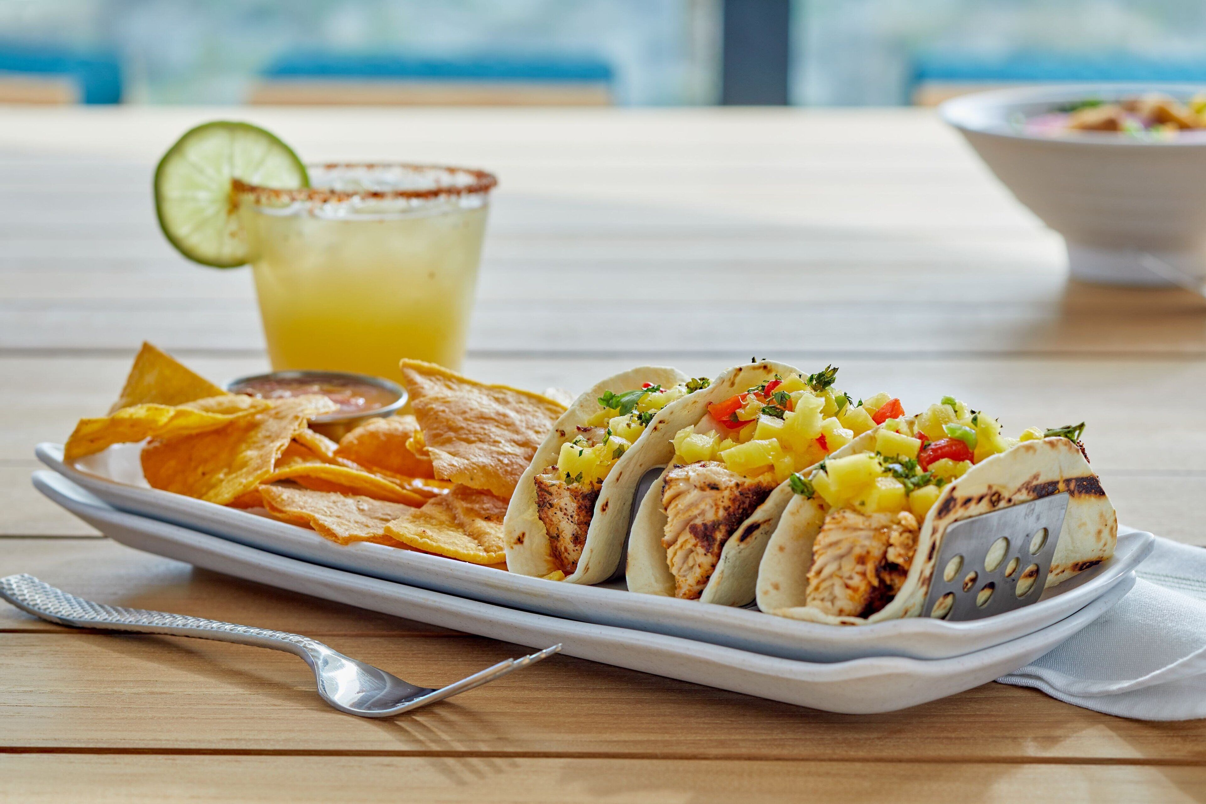 A plate of tacos with a side of chips and a drink on a wooden table.