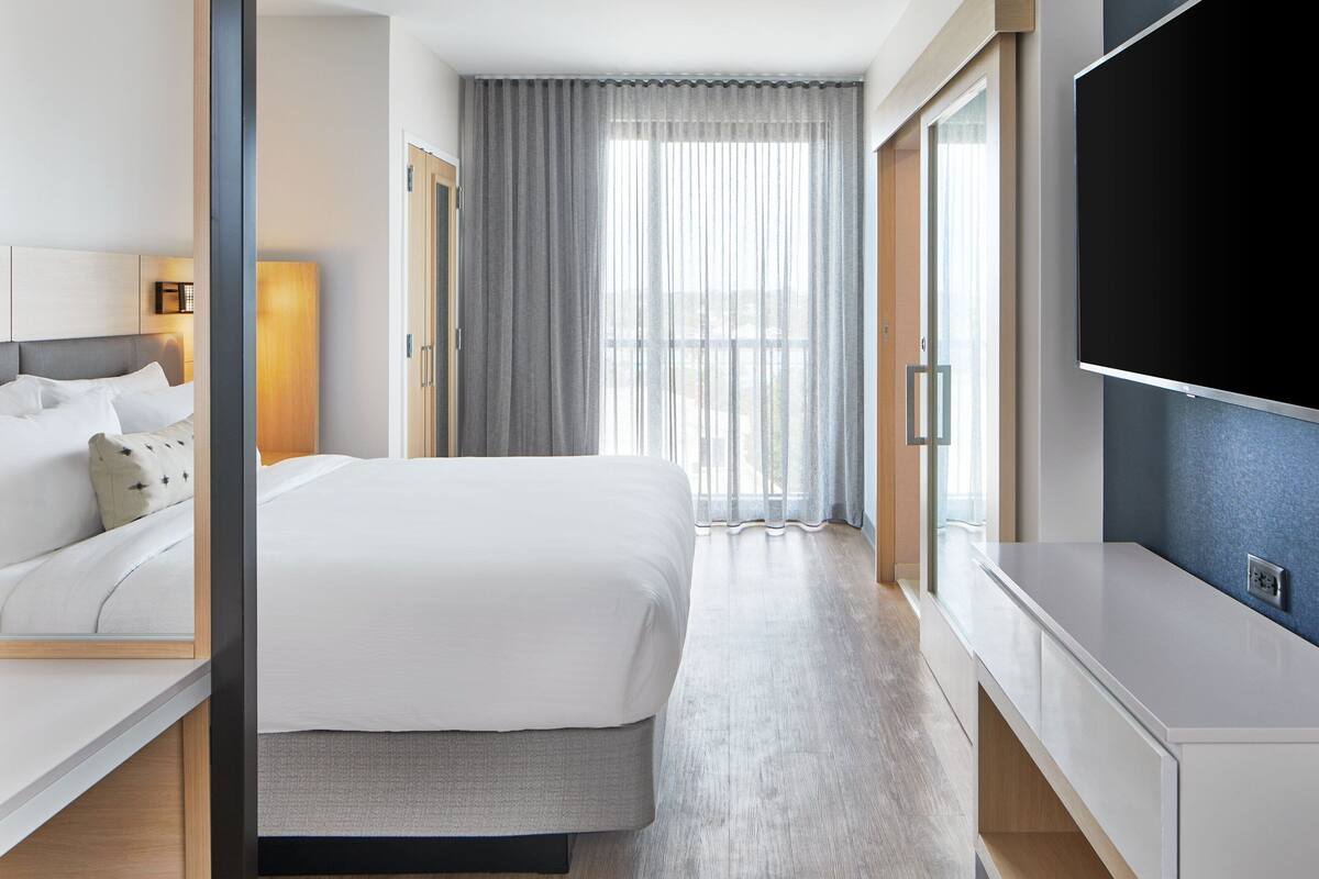 Ein Hotelzimmer mit Bett, Fernseher, Schreibtisch und einem großen Fenster mit Vorhängen.