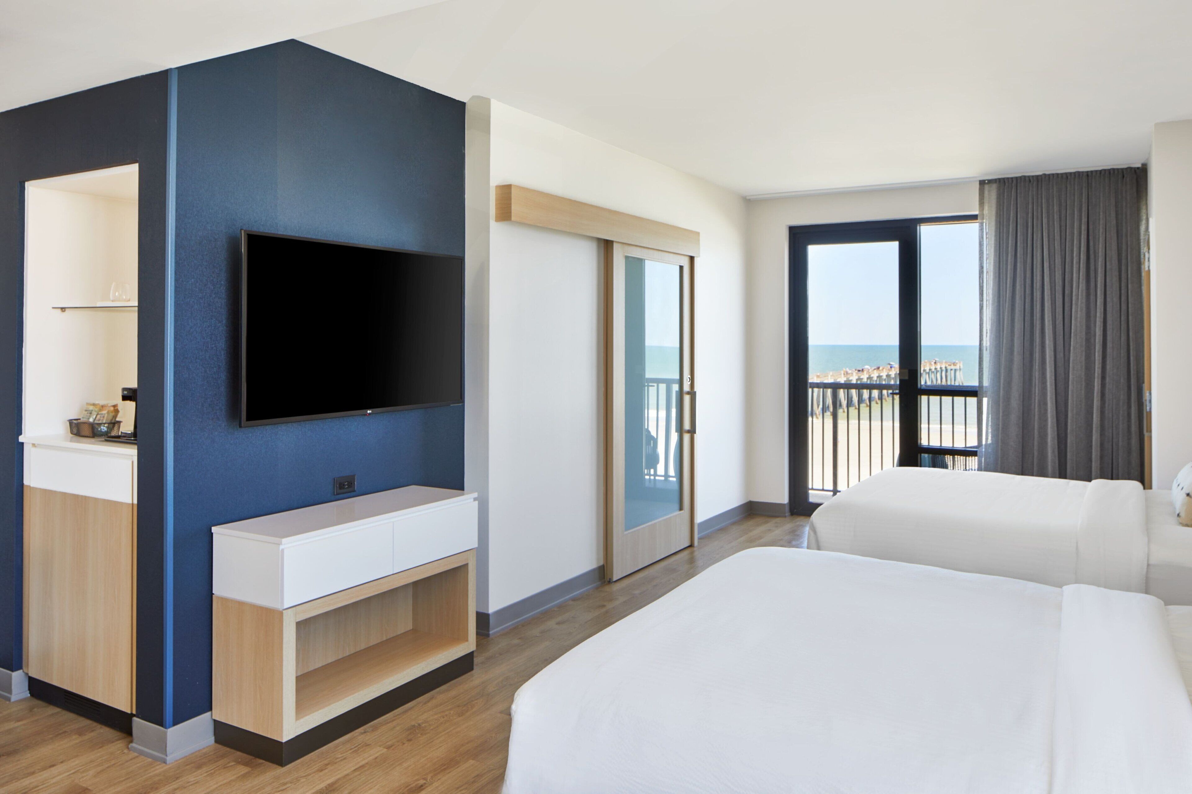 Ein Hotelzimmer mit einem großen Bett, einem an der Wand befestigten Fernseher, einem kleinen Schreibtisch und einer Schiebetür zu einem Balkon mit Meerblick.