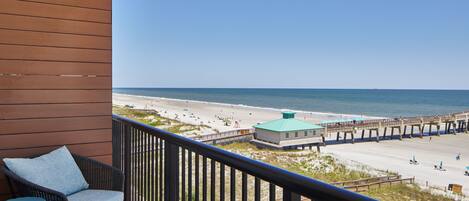 A balcony with a wicker chair and a small table, overlooking a beach with a pier and a building with a green roof.