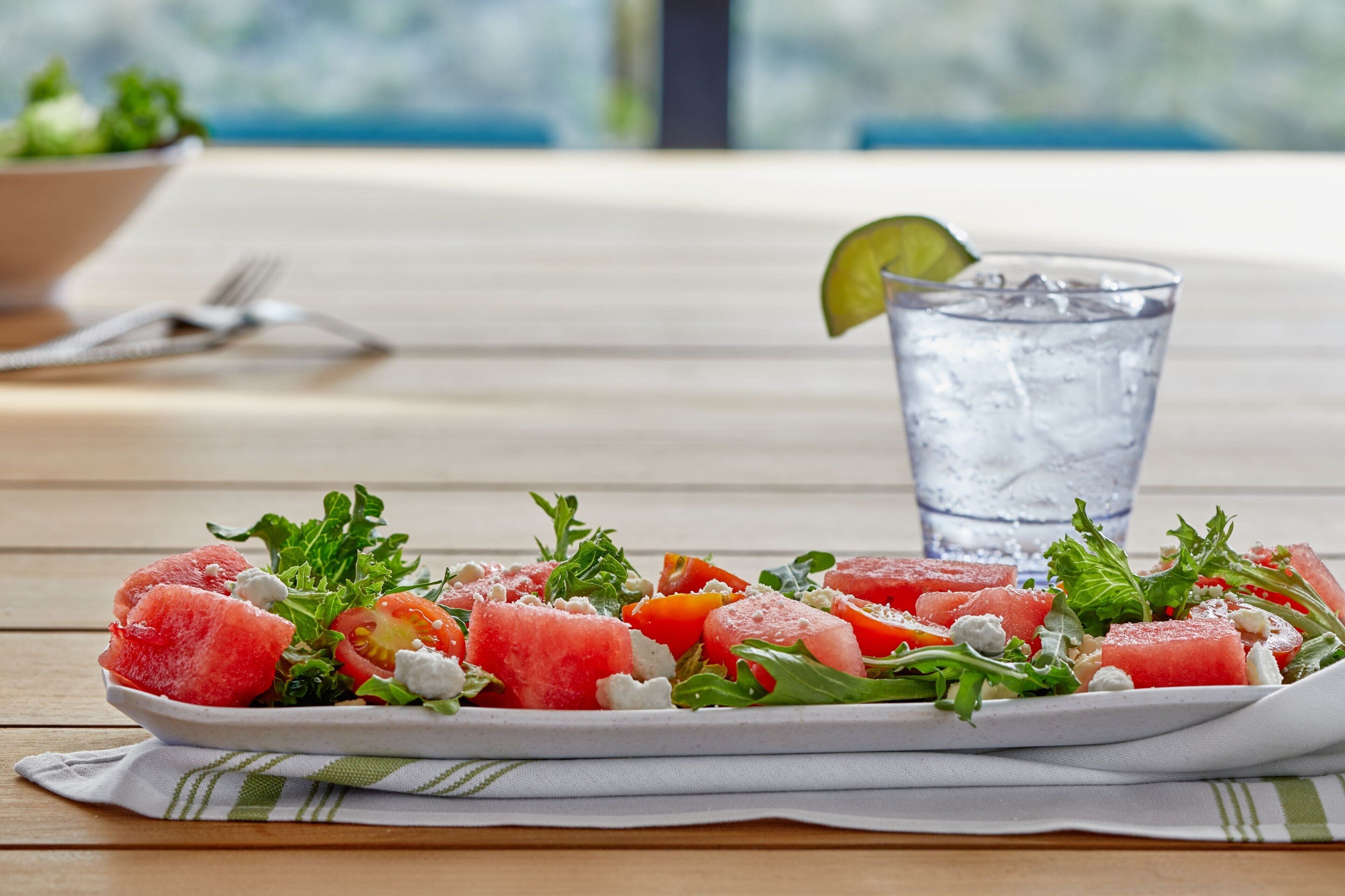 A plate of watermelon salad with feta cheese and greens, accompanied by a glass of water and a lime wedge.