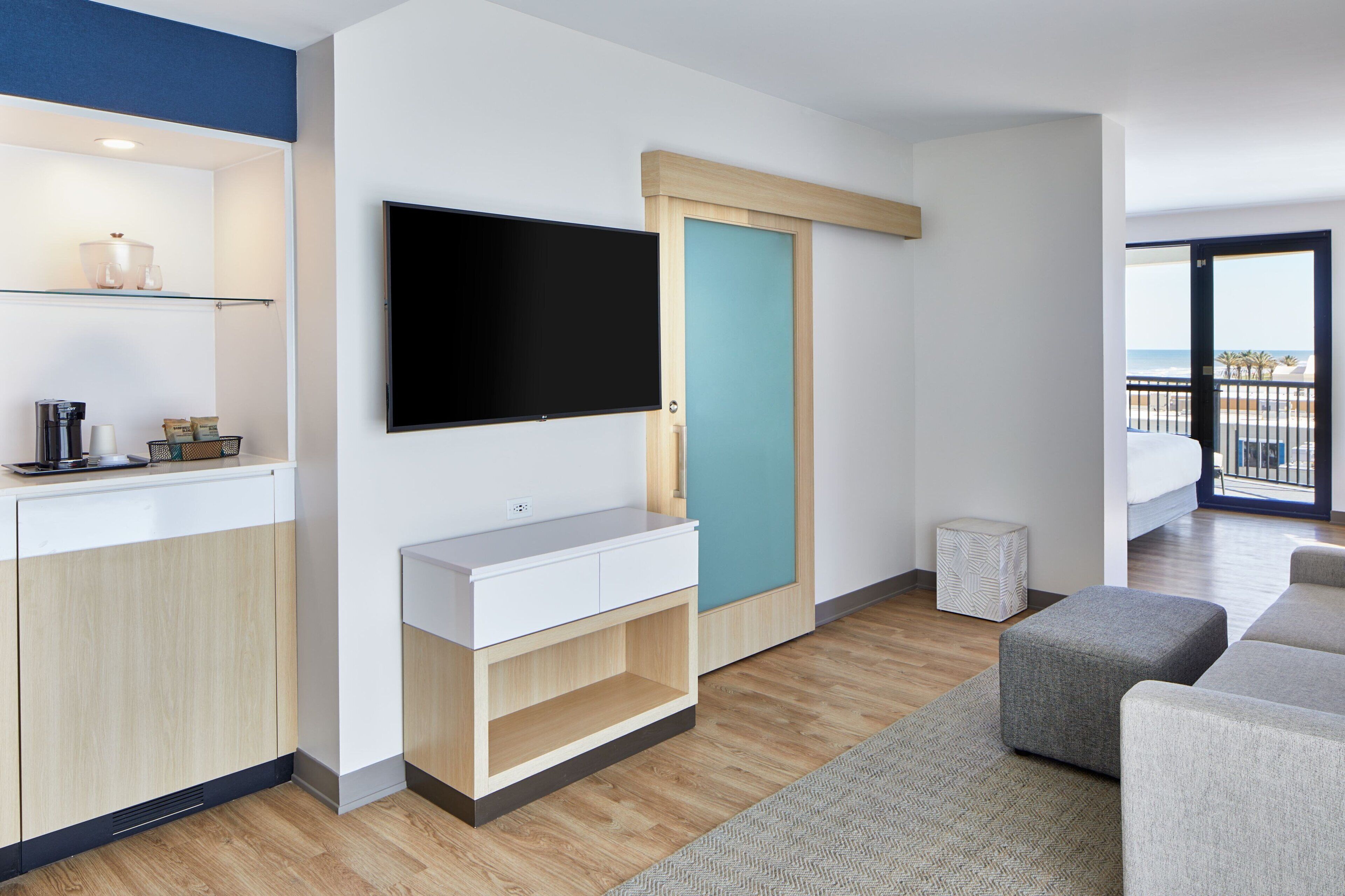 Ein modernes Wohnzimmer mit Flachbildfernseher, einer Holzkabine, einem kleinen Hocker und Blick auf den Strand durch eine Balkontür.