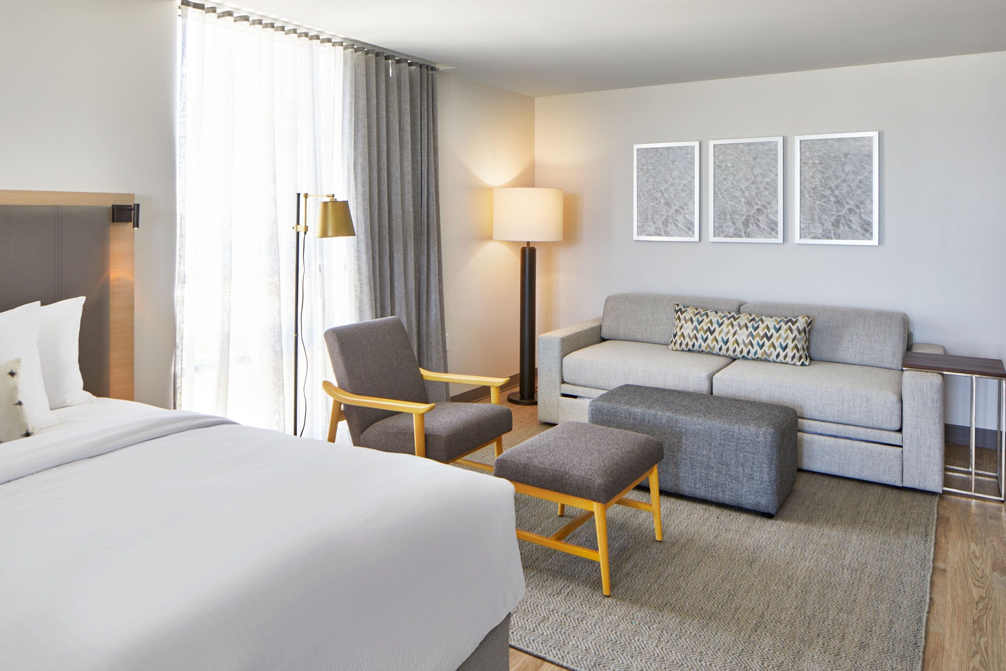 Habitació d'hotel amb un llit gran, un sofà gris, un butac armchair gris, un otomà petit, una làmpada de peu i tres quadres amb marc a la paret.