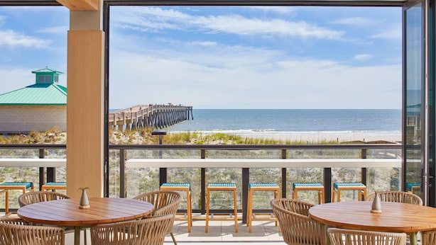 A terrace with wooden tables and chairs, overlooking a beach and a pier.