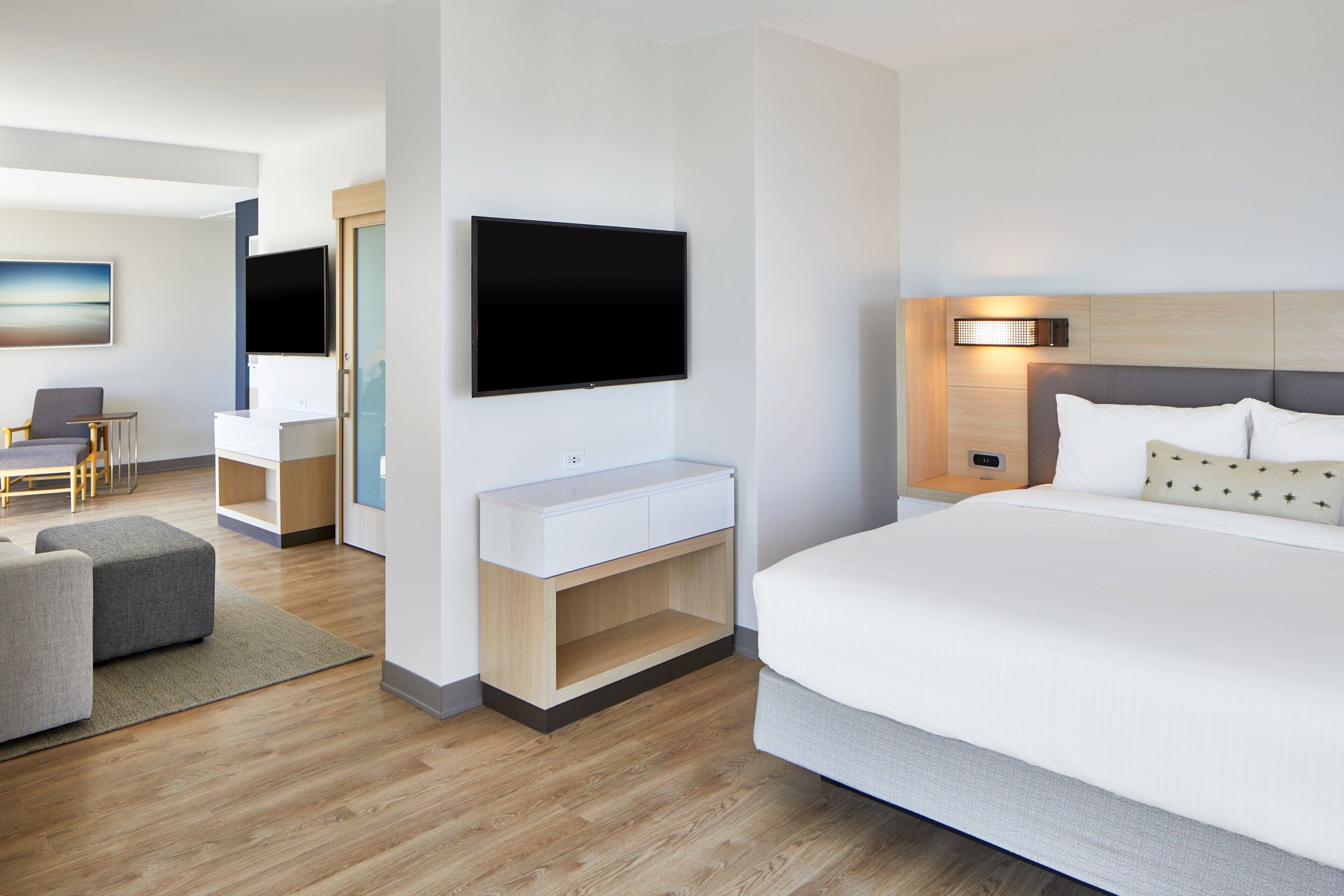 A modern hotel room with a large bed, a flat-screen TV mounted on the wall, a bedside table, and a desk with a chair.