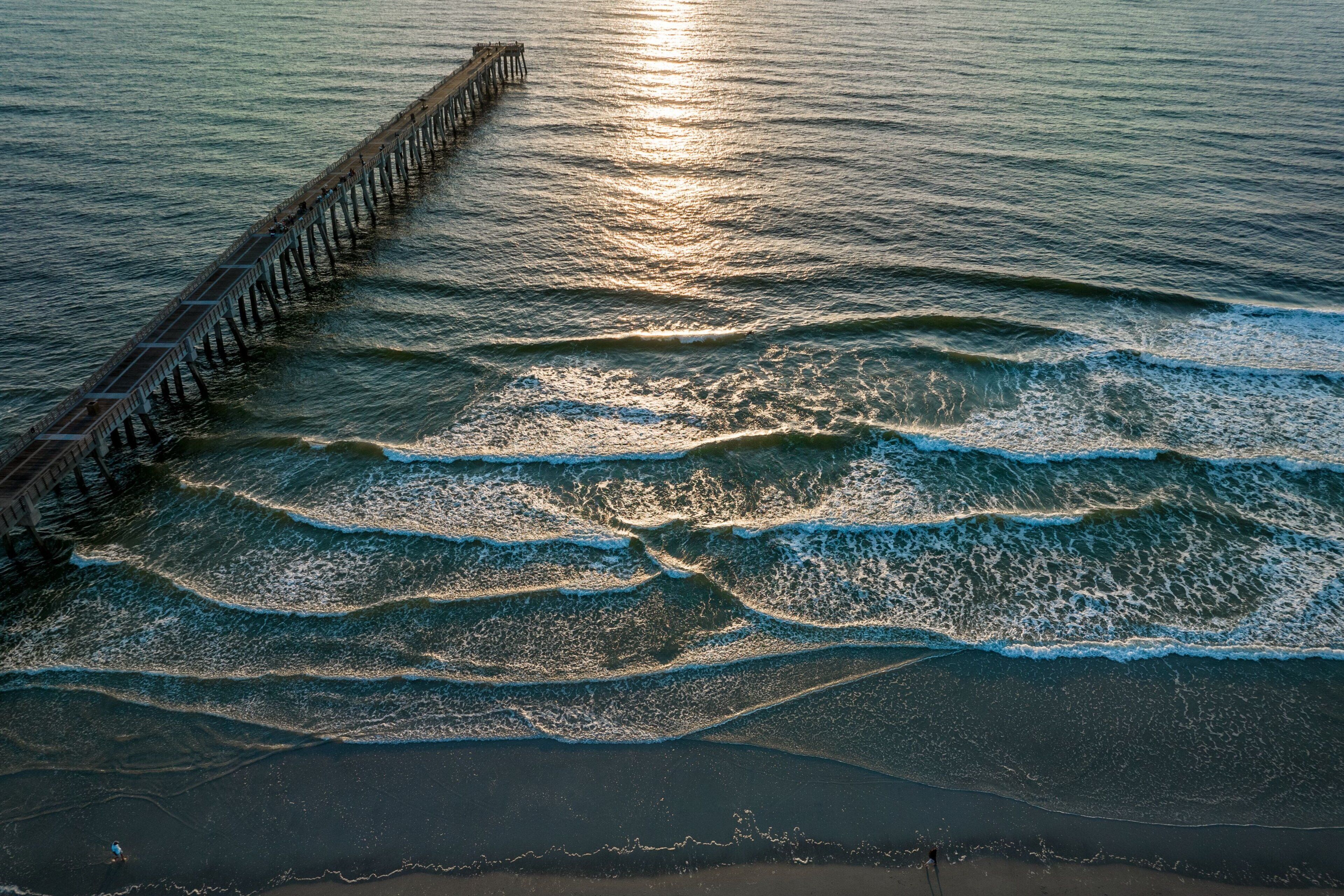 Eine Pier, die sich in ruhiges Wasser mit leichten Wellen erstreckt, ein klarer Himmel und eine entfernte Küste.