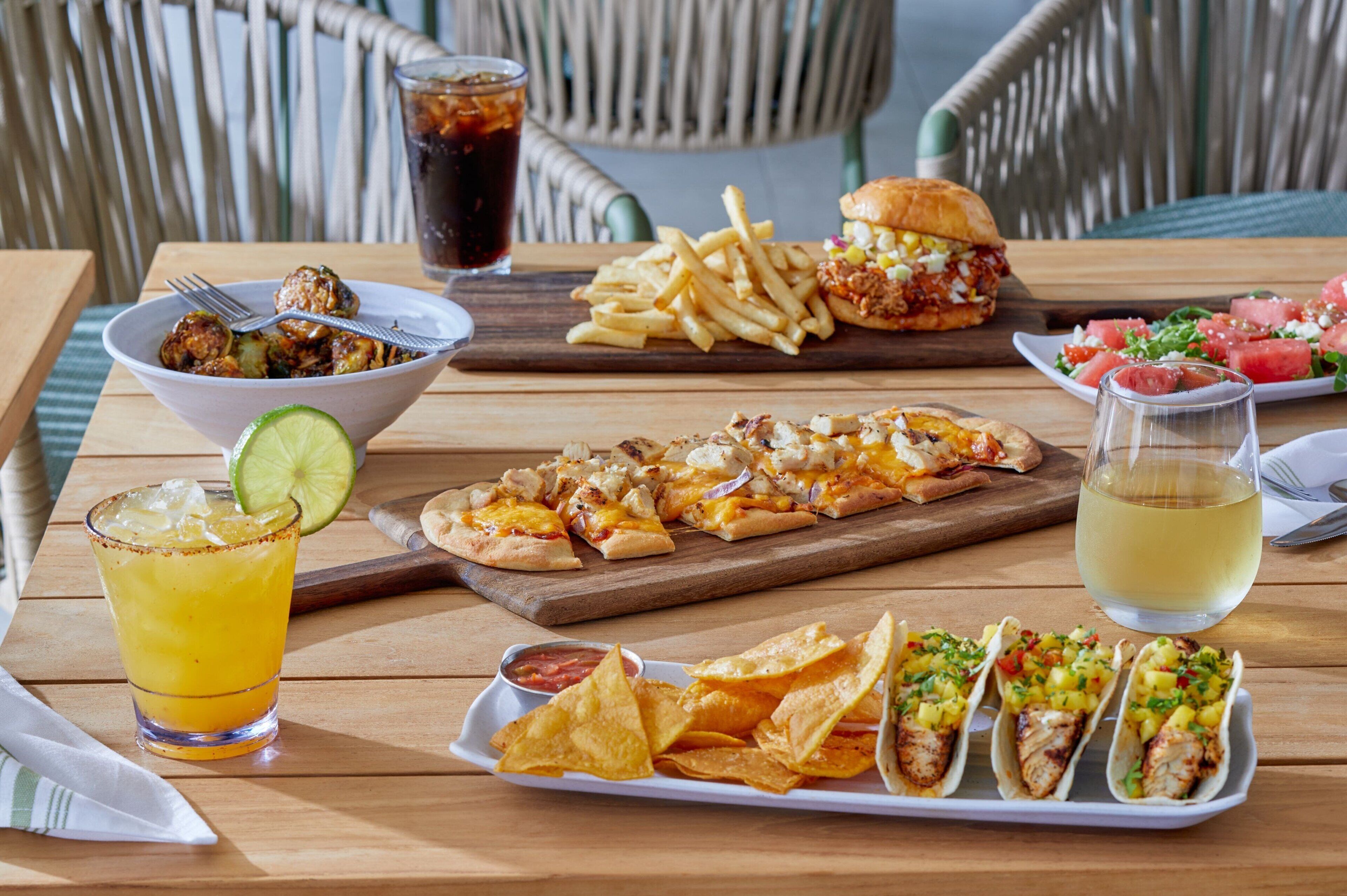Ein Holztisch mit verschiedenen Speisen, darunter ein Salat, ein Burger, eine Portion Nachos, eine Portion Tacos, ein Glas gelbliches Getränk und ein Glas dunkles Getränk.