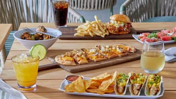 A wooden table with a variety of food items including a salad, a burger, a plate of nachos, a plate of tacos, a glass of a yellow beverage, and a glass of a dark beverage.