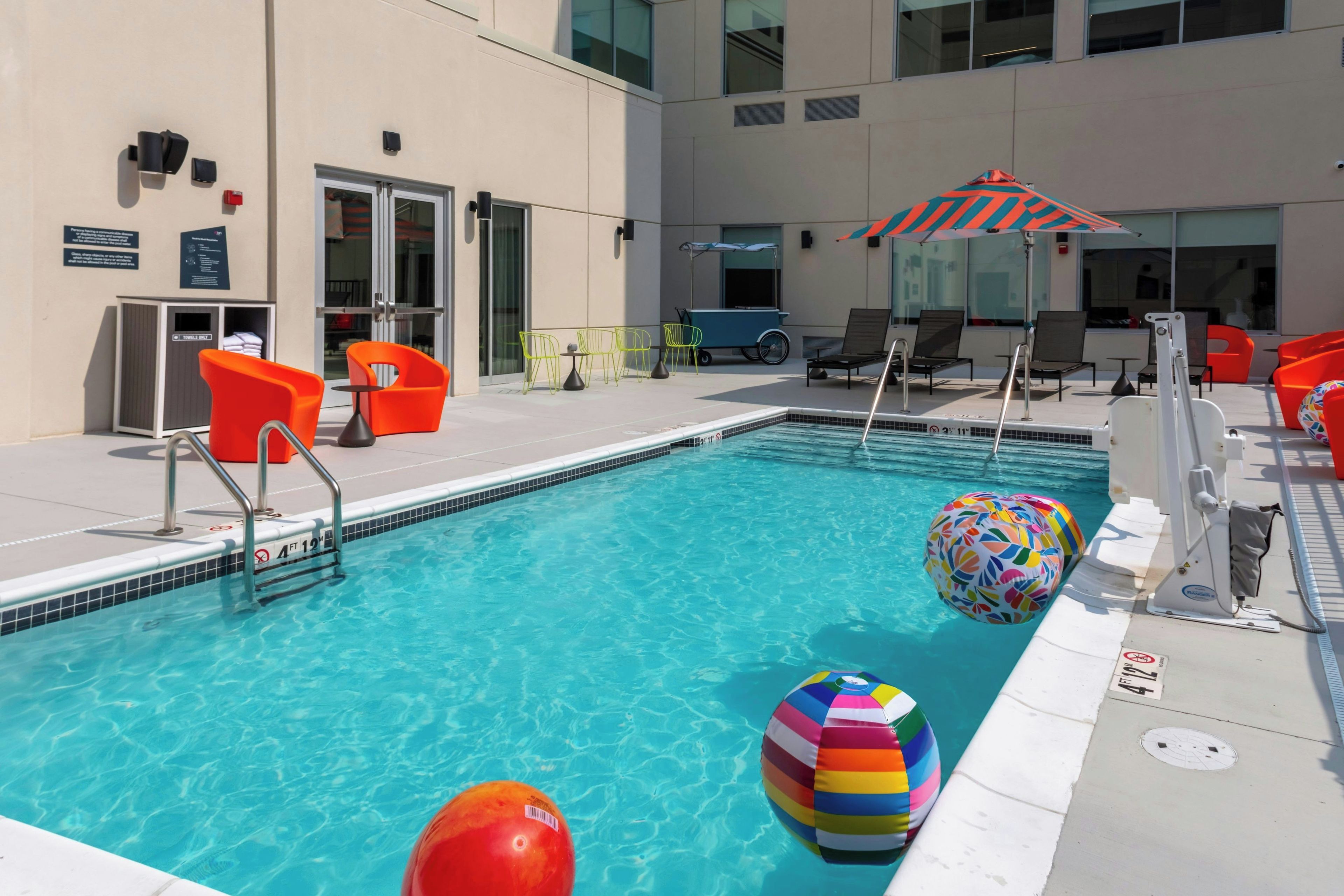 Una piscina con sillas de descanso y sombrillas, un tobogán de agua y una zona junto a la piscina con un cubo de basura y un letrero.