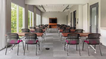 Un salón de conferencias con varias mesas y sillas, ventanas amplias en un lateral y un televisor de pantalla plana montado en la pared.