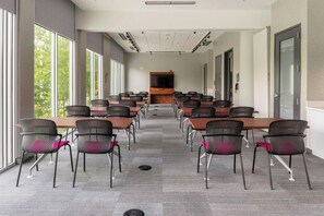 Ein Konferenzraum mit mehreren Tischen und Stühlen, gro ßen Fenstern an einer Seite und einem an der Wand befestigten Flachbildfernseher.