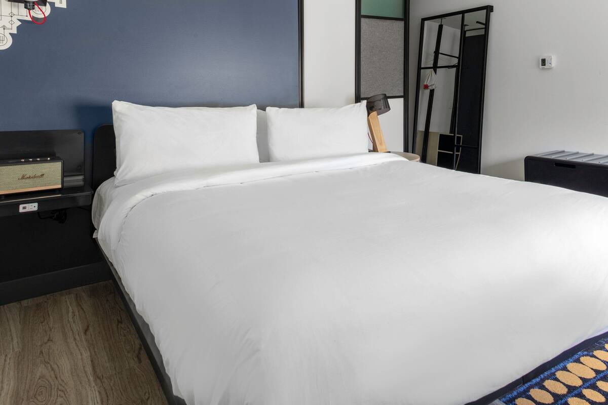 Un lit bien fait, avec du linge de lit blanc, une petite table de chevet avec une lampe et un haut-parleur fixé au mur.