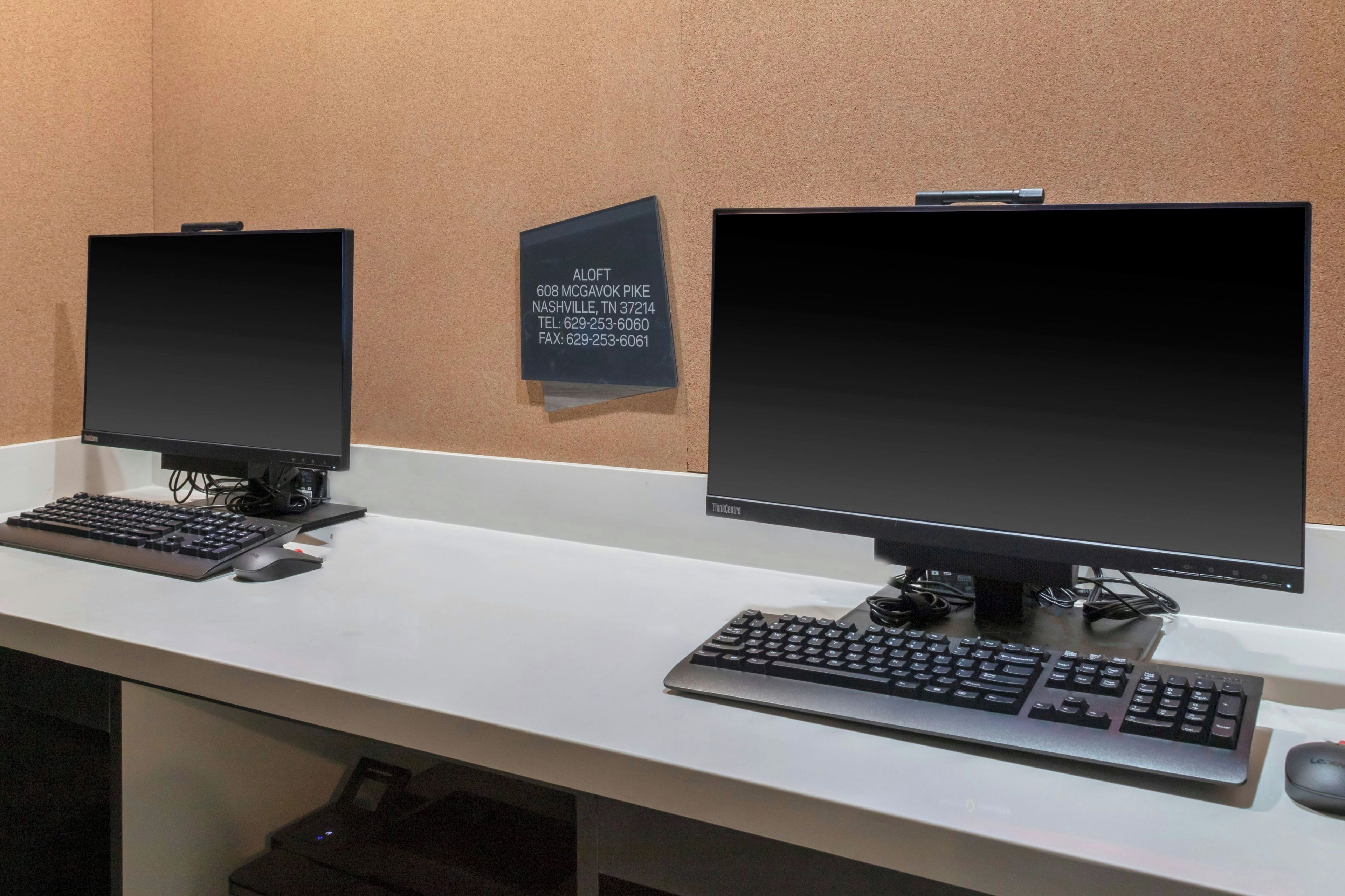 Dos monitores de ordenador sobre un escritorio blanco con teclado y ratón. Un cartel con información de contacto se ve en la pared.