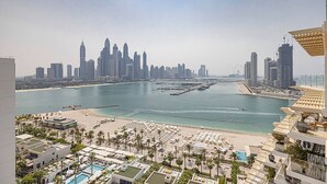 Un paisaje costero con un puerto deportivo, edificios altos y una zona de playa con sombrillas y mobiliario junto a la piscina.
