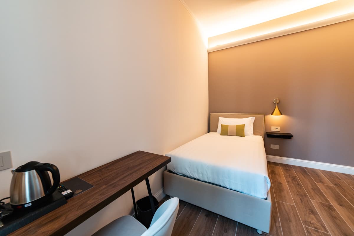 A compact hotel room with a bed, a desk, a chair, a kettle, and a bedside lamp.
