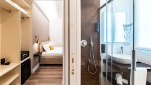 A modern hotel room with a walk-in closet, a bed with pillows, a bedside table, a small desk, a glass-enclosed shower area with a sink, and a towel rack. - Hotel del Conte (Laveno Mombello)