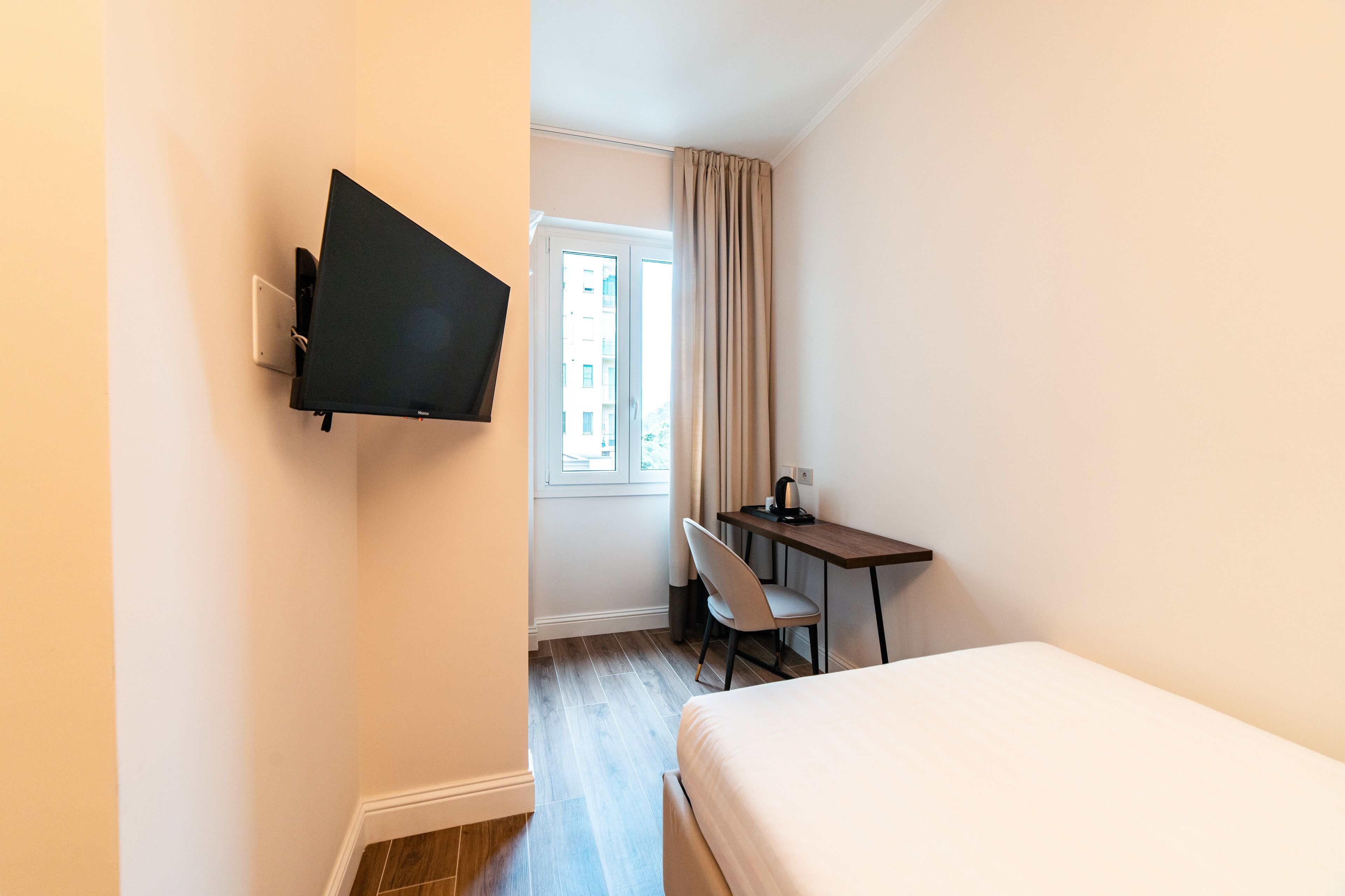 Una camera d'albergo con un letto, una scrivania con una sedia, un televisore montato a parete e una finestra con tende.