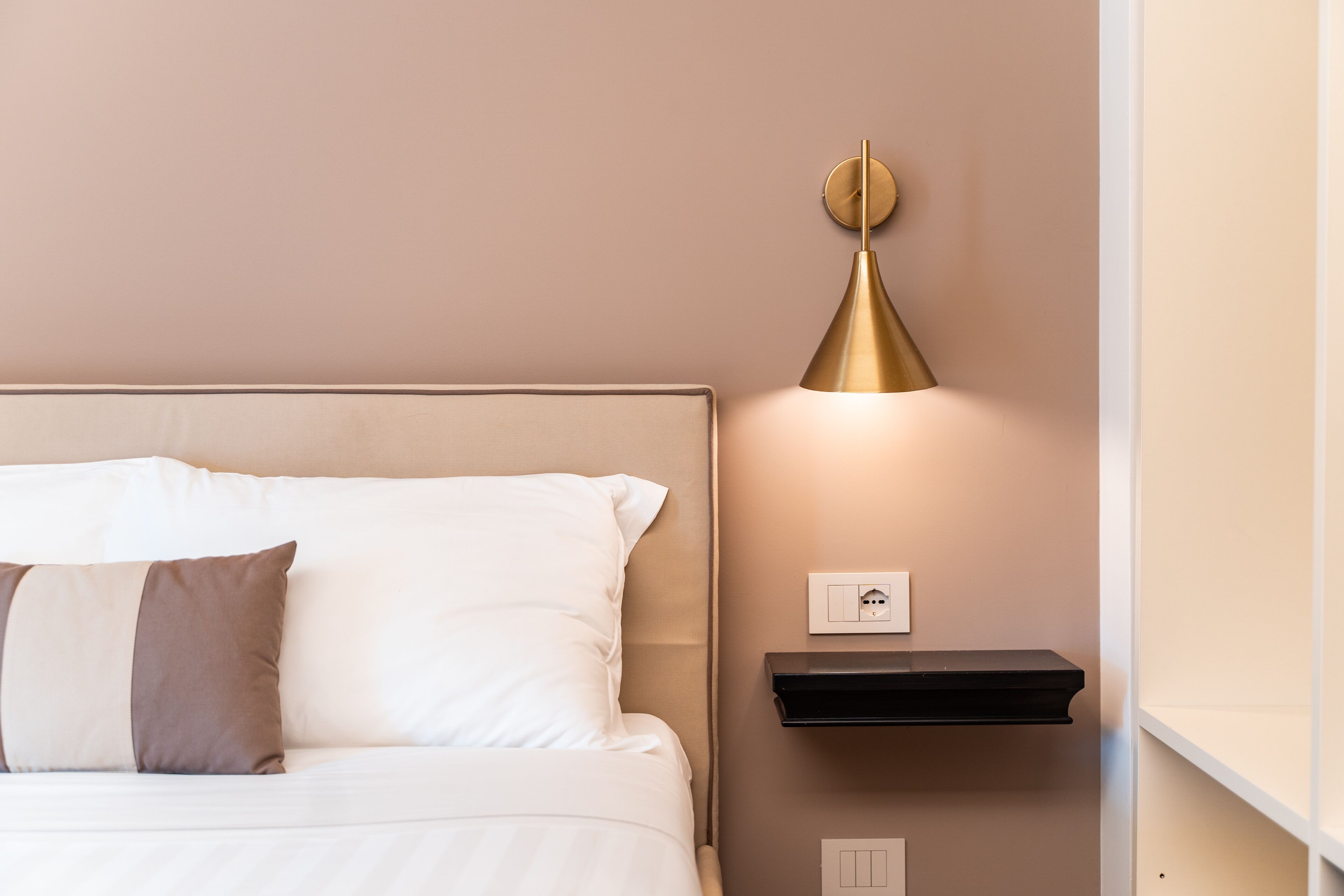 Ein ordentlich bezogenes Bett mit weißen und braunen Kissen, ein Nachttisch mit schwarzem Tablett und eine Wandlampe.