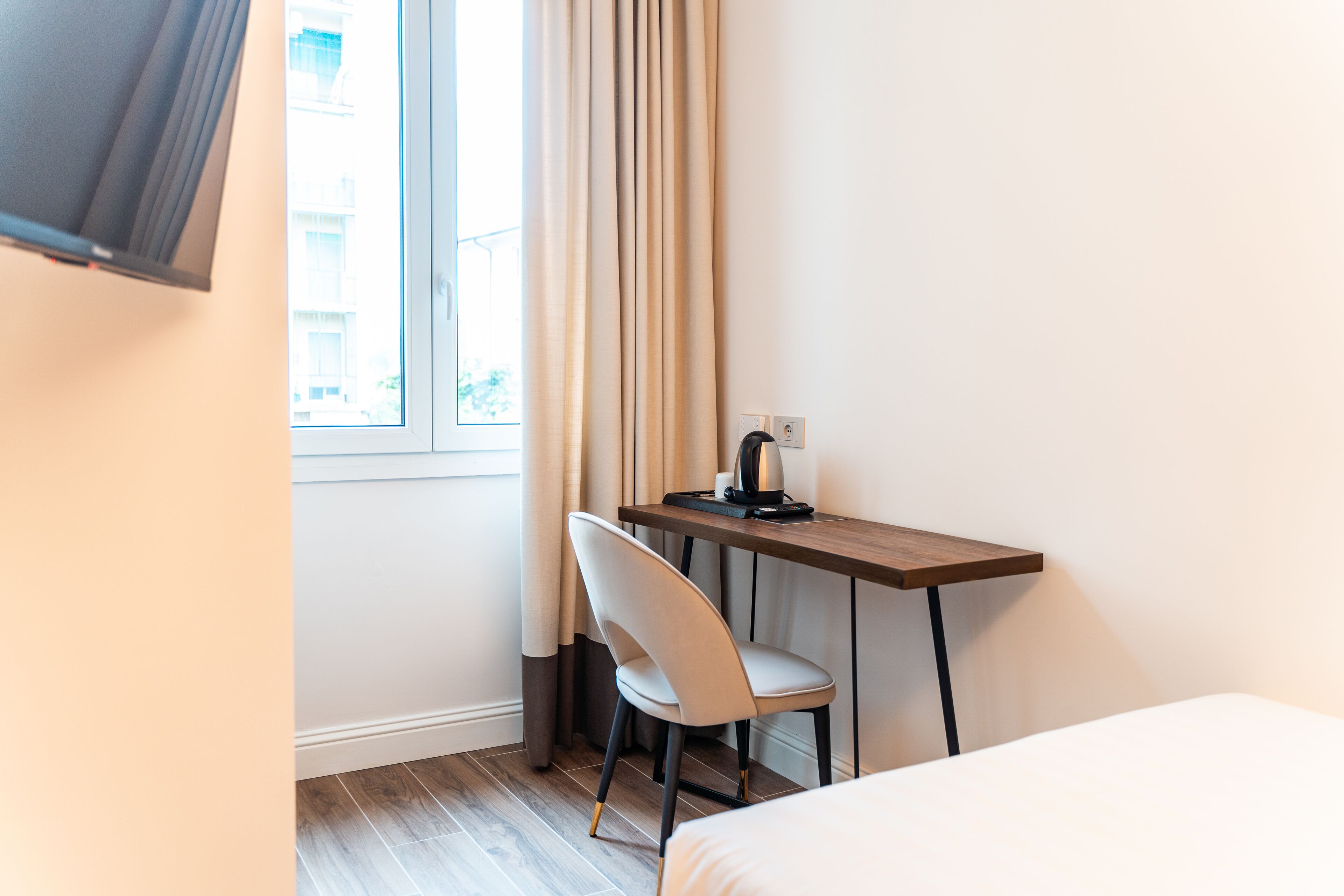 A hotel room with a bed, a desk with a chair, a window with curtains, and a television mounted on the wall.
