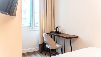 A hotel room with a bed, a desk with a chair, a window with curtains, and a television mounted on the wall.