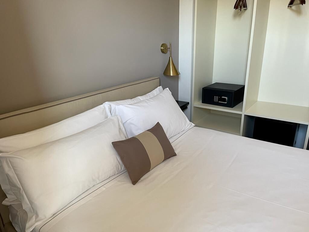 A neatly made bed with white and brown pillows, a bedside table with a black electronic device, and a gold lamp.