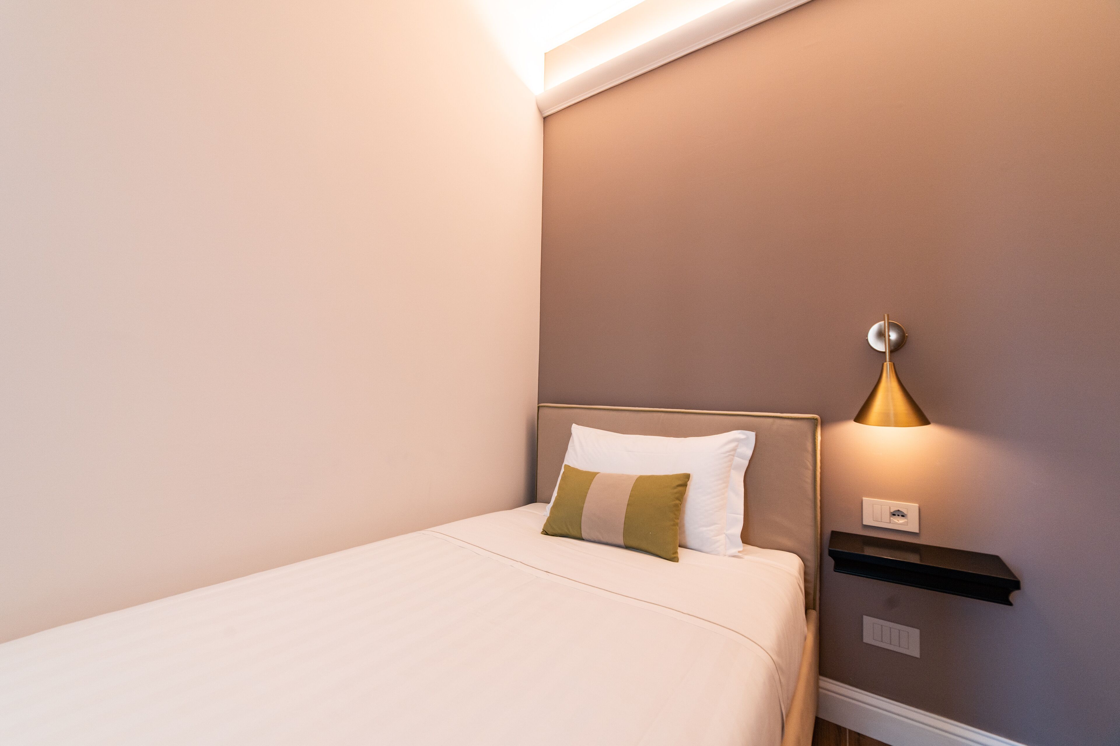 A neatly made bed with a headboard, a bedside table with a lamp, and a wall-mounted shelf.