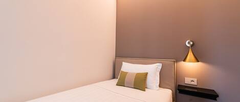 A neatly made bed with a headboard, a bedside table with a lamp, and a wall-mounted shelf.