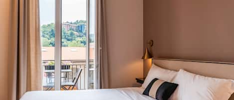 Habitación de hotel con una cama grande, una mesita de noche, una lámpara y vistas a un balcón con mobiliario exterior.