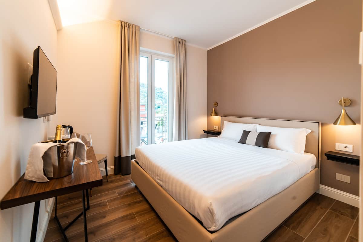 A hotel room with a large bed, a bedside table with a lamp, a flat-screen TV mounted on the wall, and a window with curtains.
