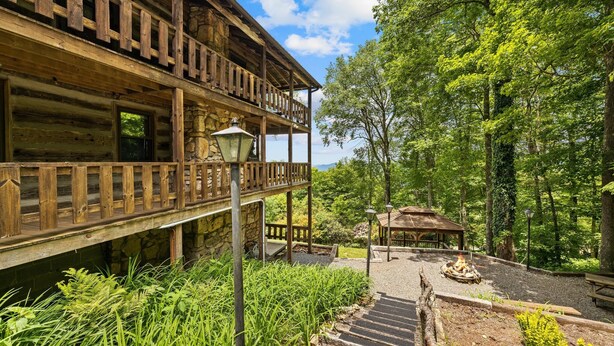A wooden cabin with a balcony, a fire pit, and a gazebo in a forested area.