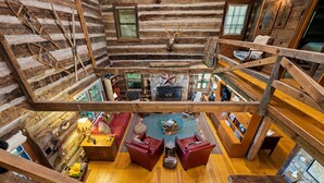 A log cabin interior with a central fireplace, red leather sofas, and a wooden staircase.