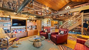 A spacious, rustic interior with wooden beams, a large flat-screen TV, leather seating, and a staircase.