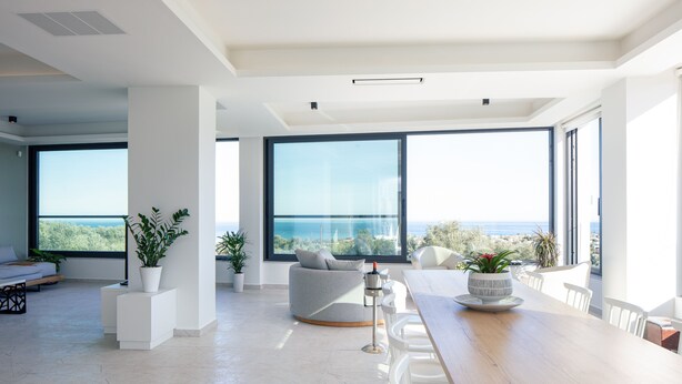 A spacious living area with a large window offering a view of the sea, a wooden dining table, and modern white furniture. - 100m2 heated pool, absolute privacy and mesmerising views! (Rethymno)